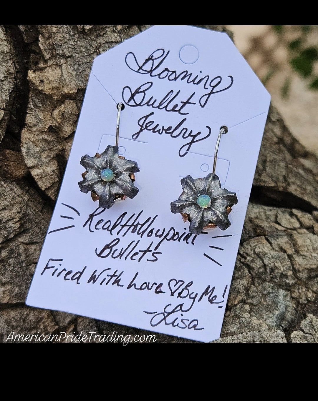 A pair of flower-shaped dangling earrings with metallic bullet details and small opal centers, displayed on a white gift tag that reads 'Bloomin Bullet Jewelry' and 'Real Hollowpoint Bullets Fired With love By Me! - Lisa.' The tag is photographed against a textured tree-bark background.
